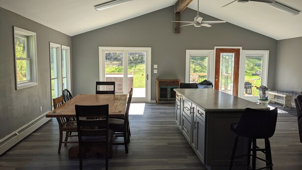 A spacious kitchen with a large island, wooden dining table, and chairs. There are multiple windows providing natural light and views of the outdoors. - large ski ranch hot tub close to killington pets ok (Mendon)