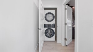 Ein Waschraum mit einer Waschmaschine und einem Trockner mit Frontlader, einem Holzboden und einer weißen Tür, die in einen Küchenbereich führt.