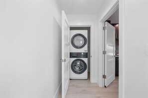 A laundry room with a front-loading washer and dryer, a wooden floor, and a white door leading to a kitchen area. - 331 | Studio | Just Steps to The Ave | King Bed | Freebee Cart |by DelrayNightly (Delray Beach)
