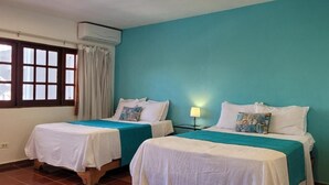 Une chambre avec deux lits, chacun recouvert d’une courtepointe blanche agrémentée de touches de couleur turquoise, une fenêtre avec des rideaux et un climatiseur fixé au mur.