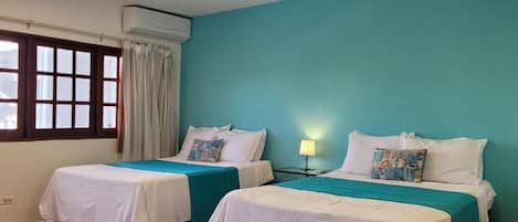A room with two beds, each with a white bedspread and teal accents, a window with curtains, and an air conditioning unit on the wall.