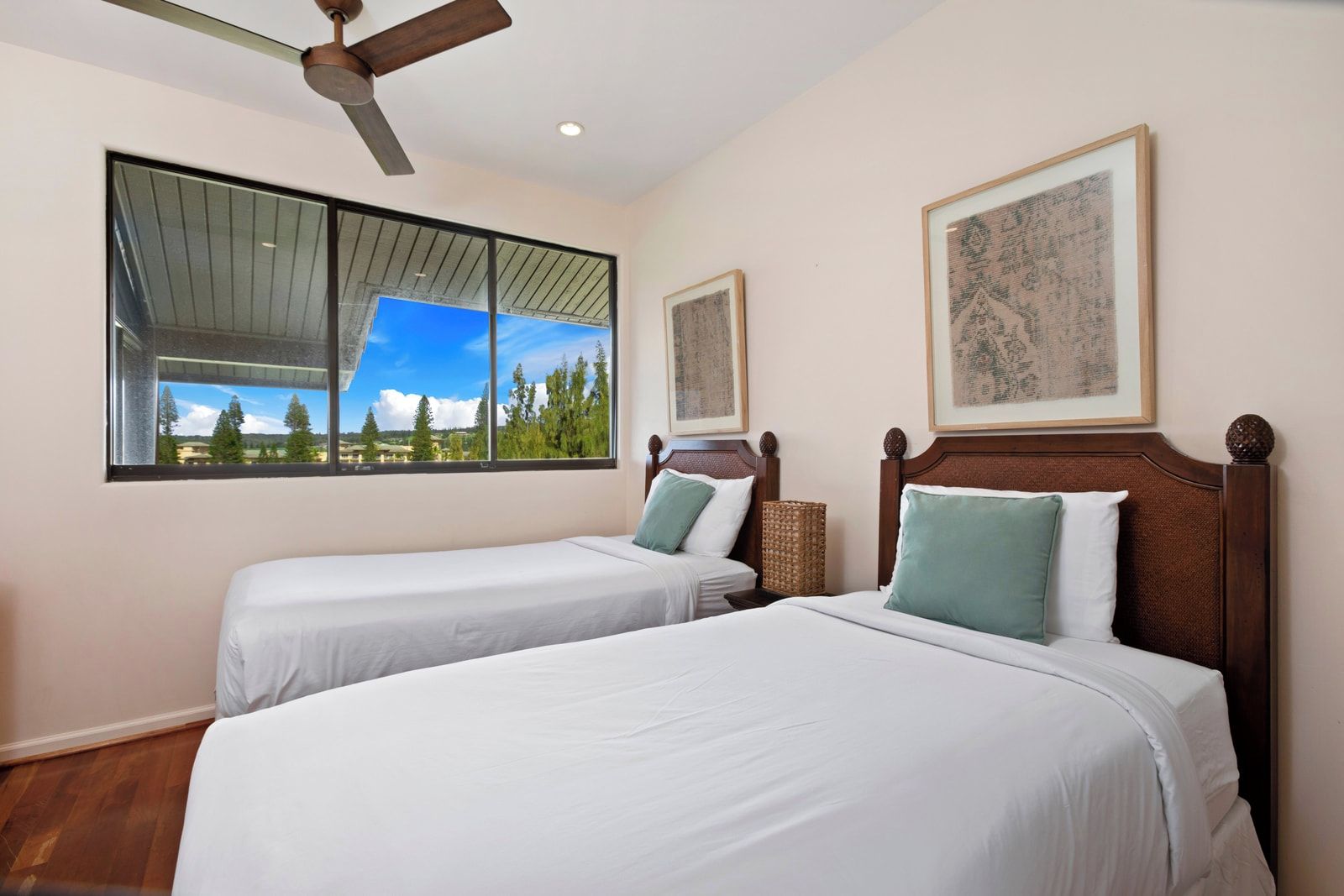 Ein Hotelzimmer mit zwei Betten, einem Deckenventilator, großen Fenstern mit Blick auf Bäume und gerahmten Kunstwerken an den Wänden.