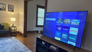 A modern living room with a large flat-screen TV on a wooden stand, a grey sofa, a small side table with a lamp, and a patterned rug. - Spacious, Family Friendly 4BR, 5Qn Beds with Garage in Columbus (Columbus)