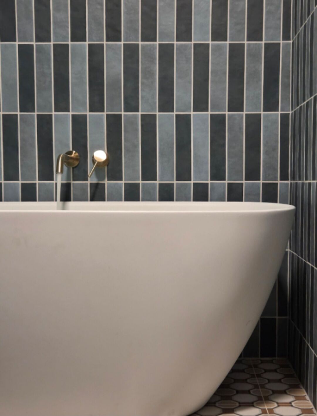 A modern bathroom with a freestanding bathtub, dark tiled walls, and a patterned floor.