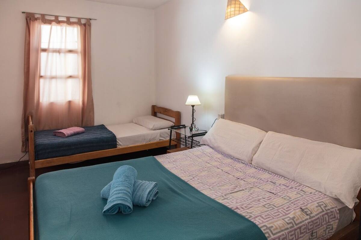 A hotel room with two beds, a bedside table with a lamp, and a window with curtains.