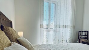 A bedroom with a bed, pillows, a nightstand, and a window with sheer curtains. - L'Ensoleillée le petit (TRASSANEL)