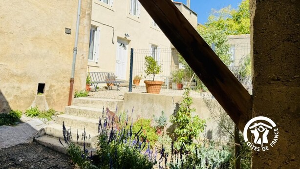 A courtyard with a stone bench, potted plants, and a building with a white door and windows. - L'Ensoleillée le petit (TRASSANEL)