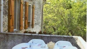 An outdoor dining area with a table and chairs, a window with wooden shutters, and a view of trees.