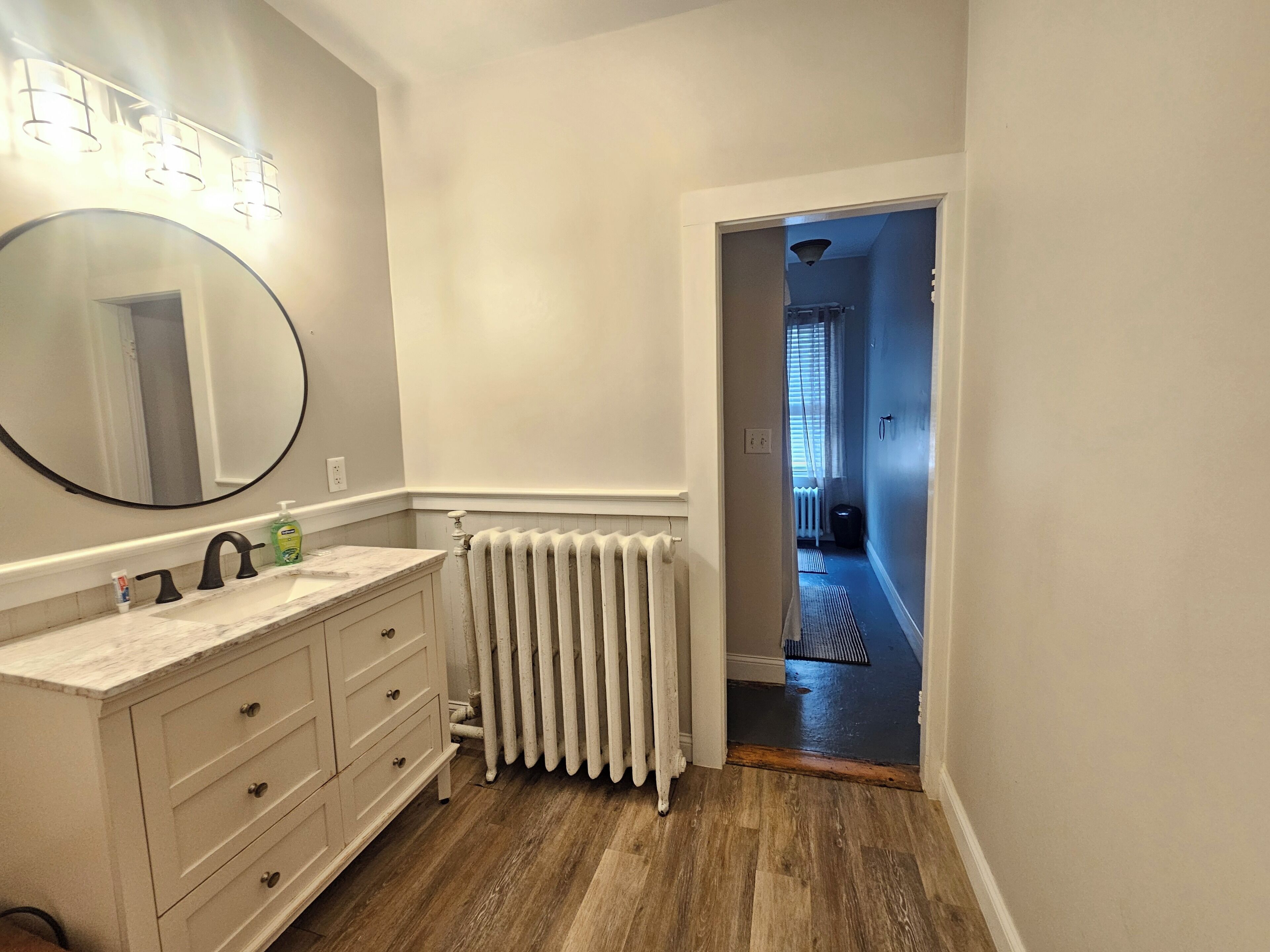 Une salle de bain avec un meuble-lavabo blanc, un miroir rond et un radiateur.