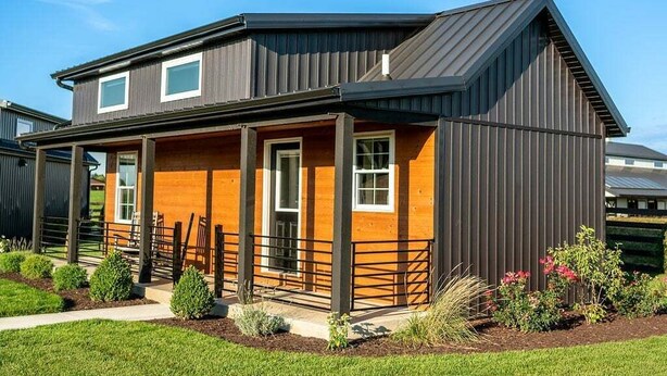 Une maison moderne à deux étages, avec une façade sombre, un porche en bois et une cour clôturée.