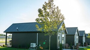 Un bâtiment avec un toit en métal et un arbre devant. Il y a des chaises et une table à l’extérieur.