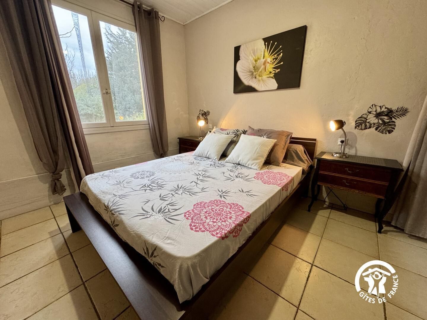 Une chambre à coucher comprenant un lit, deux tables de chevet, une commode, une fenêtre avec des rideaux et un mur orné d’un tableau représentant des motifs floraux.