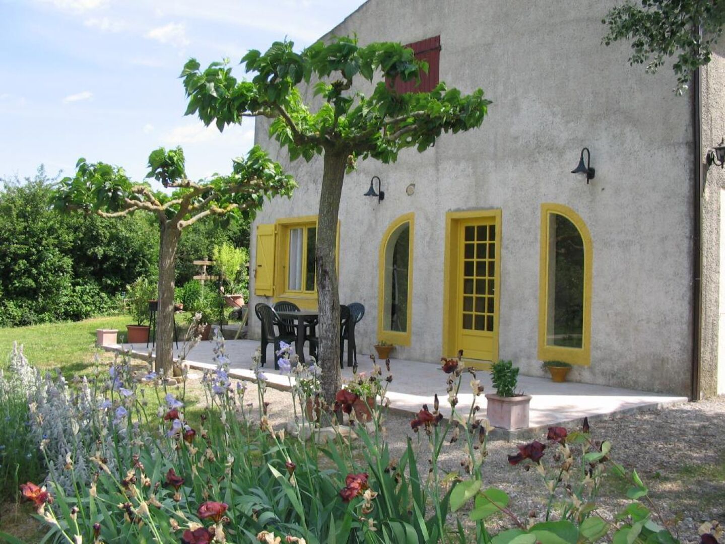 A building with yellow doors and windows, a patio with outdoor furniture, and a garden with various plants and flowers.