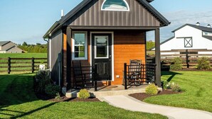 A small, single-story building with a dark roof and wooden siding, featuring a front porch with two chairs and a small table. - Simple Serenity Loft | Tiny Home #6 in Amish Country! (Shipshewana)