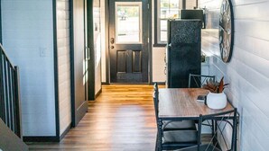 A hallway with a wooden floor, a dining table with chairs, a black refrigerator, and a door leading to another room. - Simple Serenity Loft | Tiny Home #6 in Amish Country! (Shipshewana)