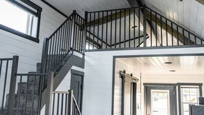 A multi-level interior with a staircase, wooden beams, and a view through an open door. - Simple Serenity Loft | Tiny Home #6 in Amish Country! (Shipshewana)