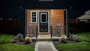 A small, single-story wooden house with a dark door and two windows, a covered porch with chairs, and a paved pathway leading to the entrance. - Simple Serenity Loft | Tiny Home #6 in Amish Country! (Shipshewana)