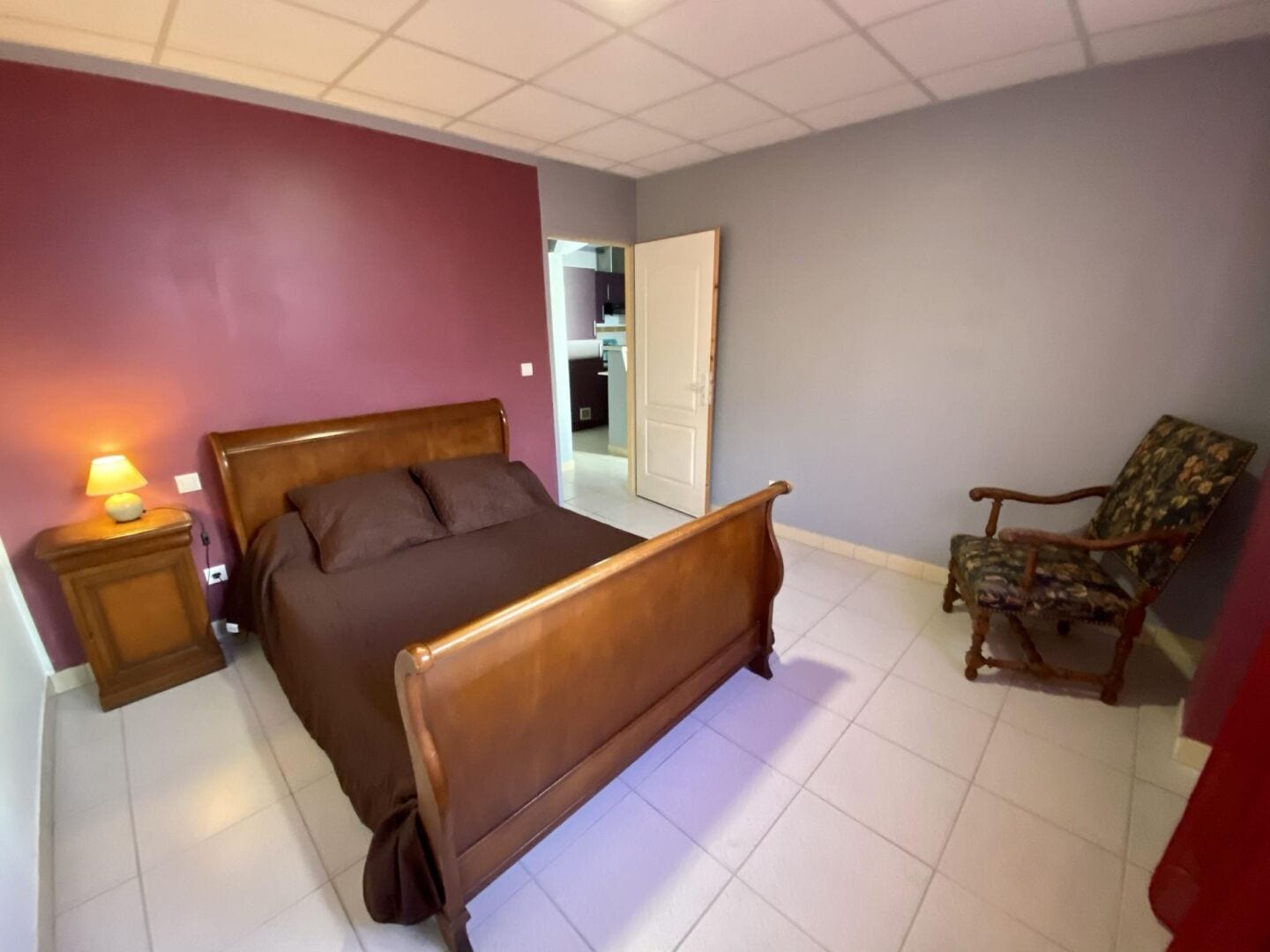 A bedroom with a wooden bed, a nightstand with a lamp, a chair, and a door leading to another room.