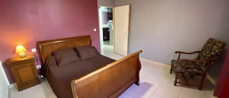 A bedroom with a wooden bed, a nightstand with a lamp, a chair, and a door leading to another room.