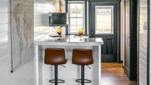 A kitchen with a central island, two bar stools, and a world map on the wall. - Whispering Cottage #3 | Shewana Tiny Home Village (Shipshewana)