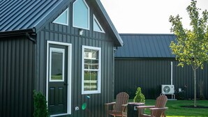A modern house with a dark exterior, a large front door, and a covered patio area with two wooden chairs and a small table. - Whispering Cottage #3 | Shewana Tiny Home Village (Shipshewana)