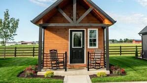 A small wooden cabin with a black door, two windows, and a porch with two chairs. - Whispering Cottage #3 | Shewana Tiny Home Village (Shipshewana)