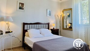 Una cama bien hecha con sábanas blancas, un cabecero de madera y una manta gris. Se observa una mesita de noche con una lámpara, un adorno pequeño y un cuadro en la pared. Un espejo grande refleja parte de la habitación, y se ve una ventana con cortinas traslúcidas.