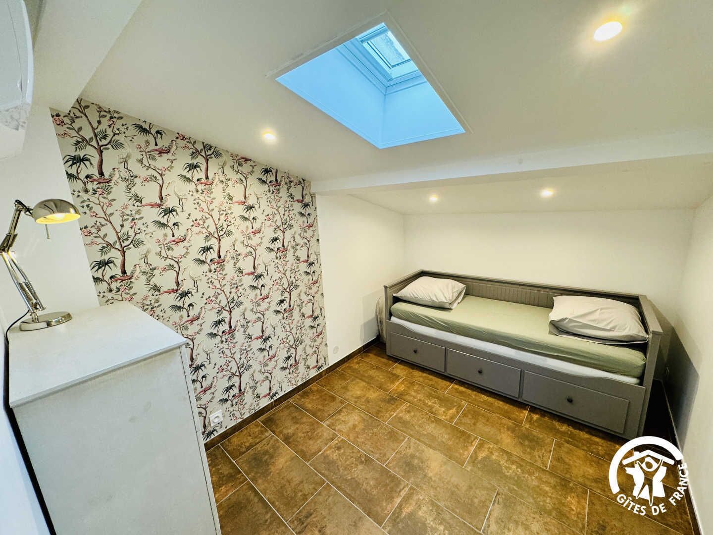 Una habitación compacta con tragaluz, una cama, un escritorio con lámpara y un papel tapiz estampado.