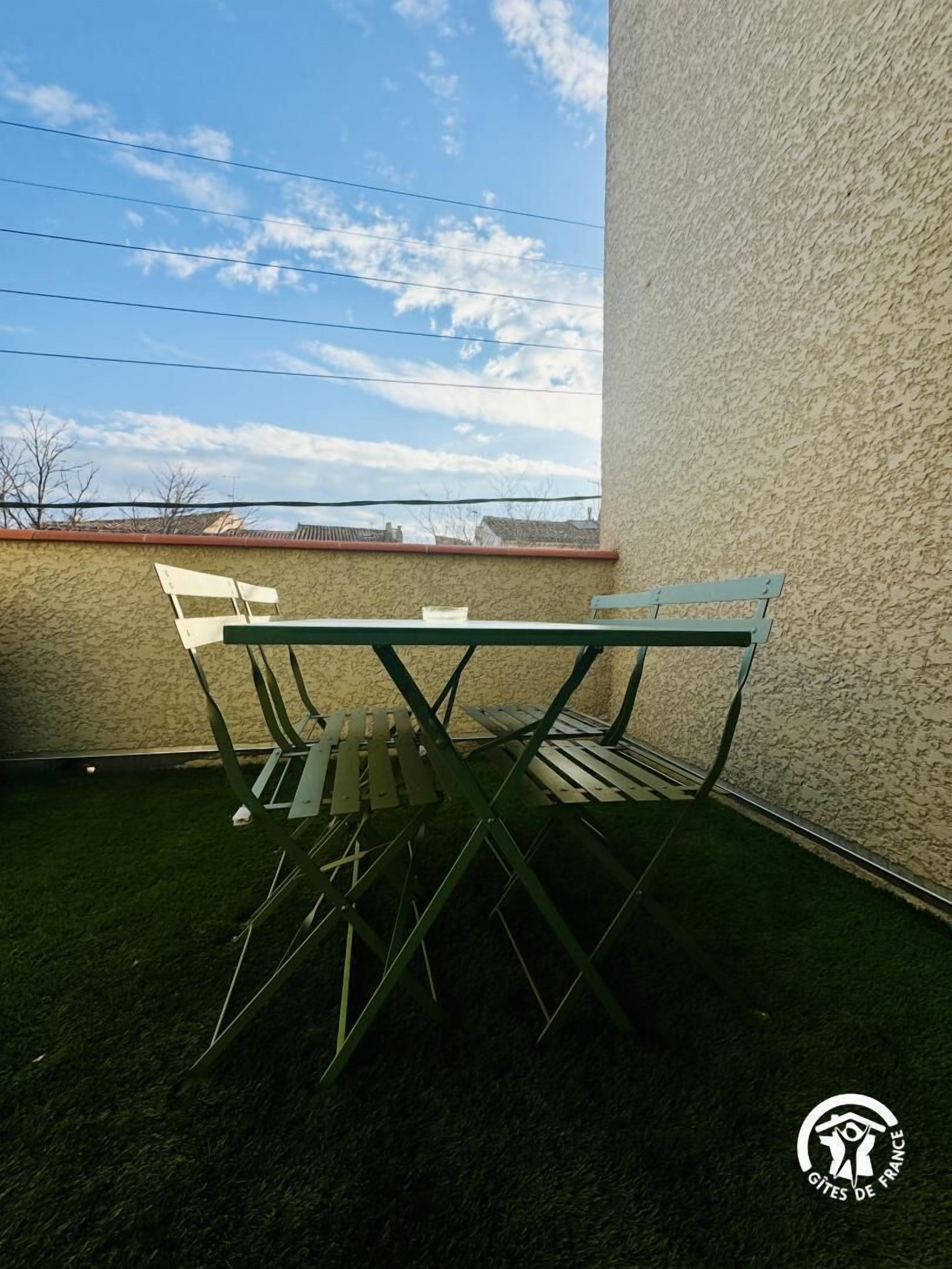 A small outdoor patio area with a metal table and chairs, surrounded by a textured wall and a grassy area.