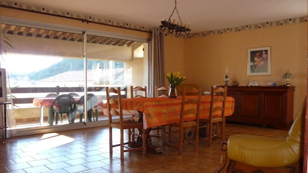 A dining area with a table, chairs, and a yellow chair. There is a window with a view of a building and a mountain. - La Loggia - ROQUEFORT DES CORBIERES (ROQUEFORT DES CORBIERES)