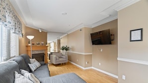 Ein Wohnzimmer mit grauem Ecksofa, Kamin, Wandfernseher und einem zur anderen Wohnung führenden Gang.