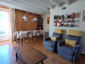A cozy living room with a wooden dining table, two armchairs, a bench, and a bookshelf with various items.