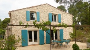 Una casa de piedra de dos pisos con contraventanas azules, un patio adoquinado y una pérgola cubierta de enredaderas.