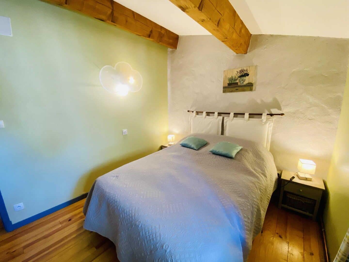 A bedroom with a bed, two pillows, a nightstand with a lamp, a wall-mounted picture, and a wooden ceiling beam.