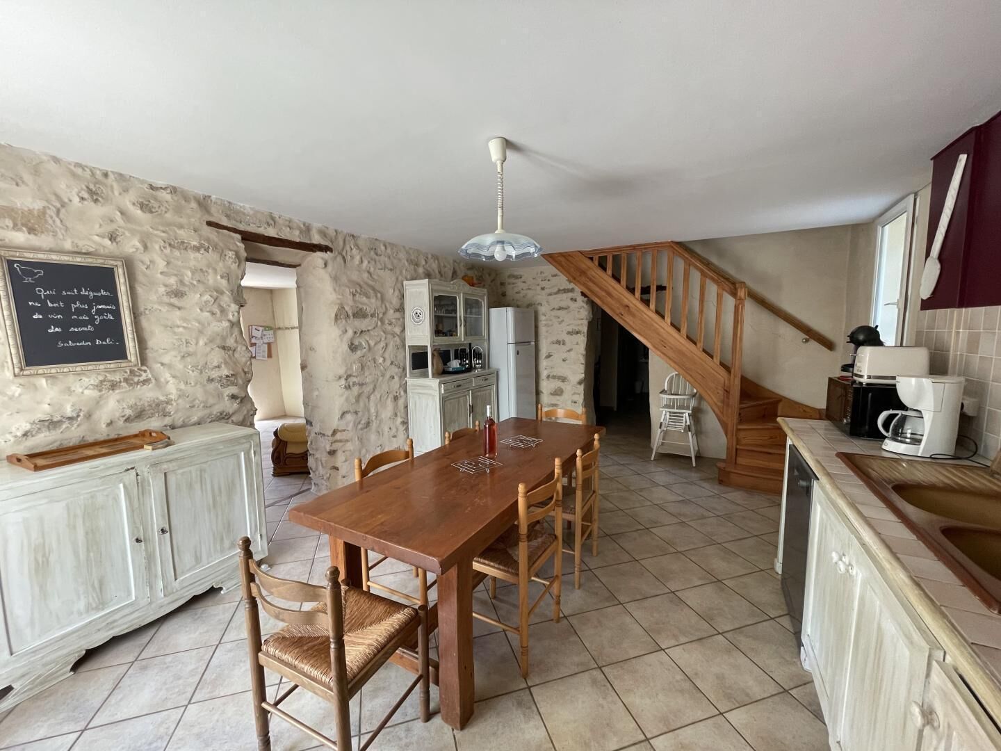 A rustic kitchen with a wooden dining table, white cabinets, and a staircase leading to another level.