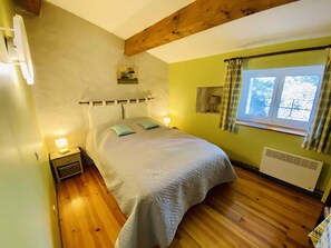 A bedroom with a bed, bedside lamps, a window with curtains, a radiator, and a wooden ceiling beam. - Le Camberaud - MAILHAC (MAILHAC)