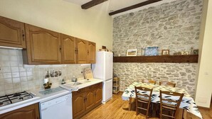 Une cuisine avec des meubles en bois, un réfrigérateur blanc, une cuisinière et un évier. On y trouve une table à manger avec des chaises, et un mur en pierre en arrière-plan.