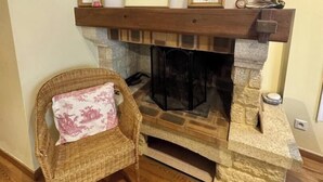 Une cheminée avec un manteau en pierre, un fauteuil en osier agrémenté d’un coussin rose, et trois tableaux encadrés au-dessus du manteau.