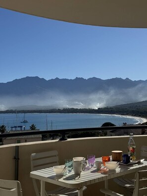 A balcony with a table set for dining, overlooking a coastal view with mountains and a body of water. - Terrasse vue mer et montagne – Calvi centre et plage (CALVI)