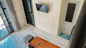 Une chambre d’hôtel moderne avec un sol en marbre, une table en bois, des sièges rembourrés de couleur grise, un téléviseur à écran plat fixé au mur et une vue sur la ville grâce à de grandes fenêtres.