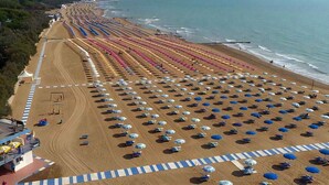 Uma praia com fileiras de guarda-sóis e espreguiçadeiras, um calçadão de areia e vista para o oceano.
