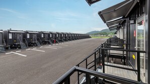 A row of portable toilets with numbered stalls, a paved area, and a building with a roofed walkway. - HOTEL R9 The Yard Tagawakawara (Kawara)