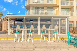 Un espacio al aire libre cubierto con mesas y sillas, vista a la piscina y un edificio con balcones.