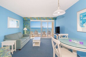 Ein Zimmer mit einer Couch, einem Tisch, Stühlen, einem Fernseher und einem Balkon mit Meerblick.