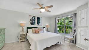 Une chambre à coucher comprenant un lit, des tables de chevet, une chaise, un ventilateur de plafond et une fenêtre avec des rideaux.