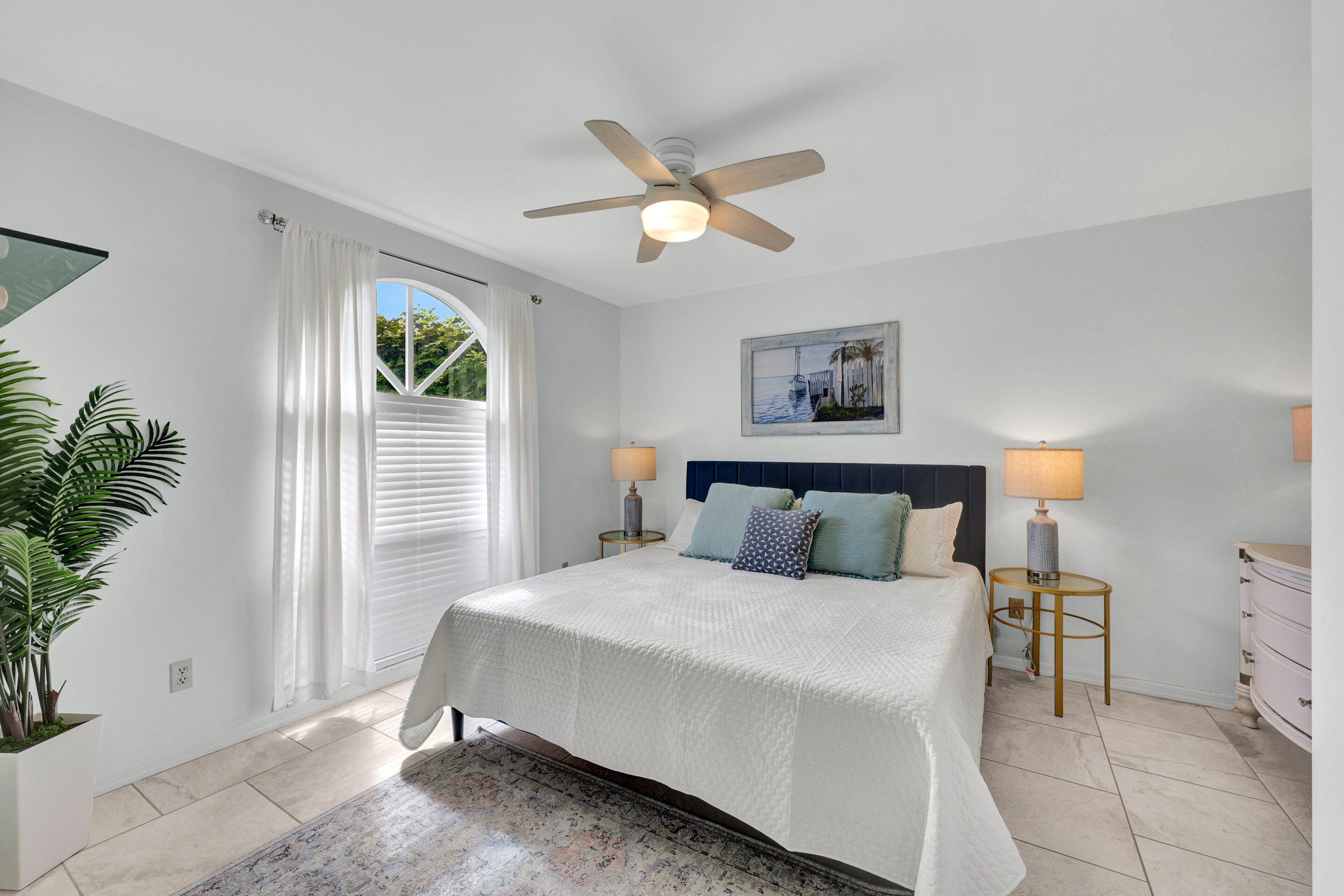 Un dormitori amb un llit, ventilador de sostre, dues taules de nit amb làmpades, una petita taula auxiliar, un armari i una finestra amb cortines, i una planta en test.