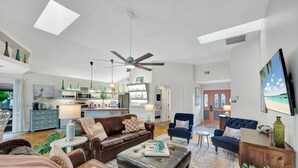 Un vaste salon avec un canapé en cuir marron, un fauteuil bleu et une table basse. On aperçoit au fond un ventilateur de plafond, un téléviseur et un coin cuisine.