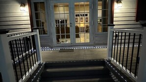 Escadaria que leva a um deck de madeira com porta de vidro, guarda-corpos brancos e iluminação externa.