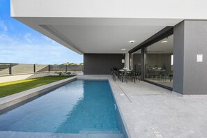 Ein moderner Poolbereich im Freien mit Esstisch und Stühlen, einem Grill und Blick auf den Himmel.