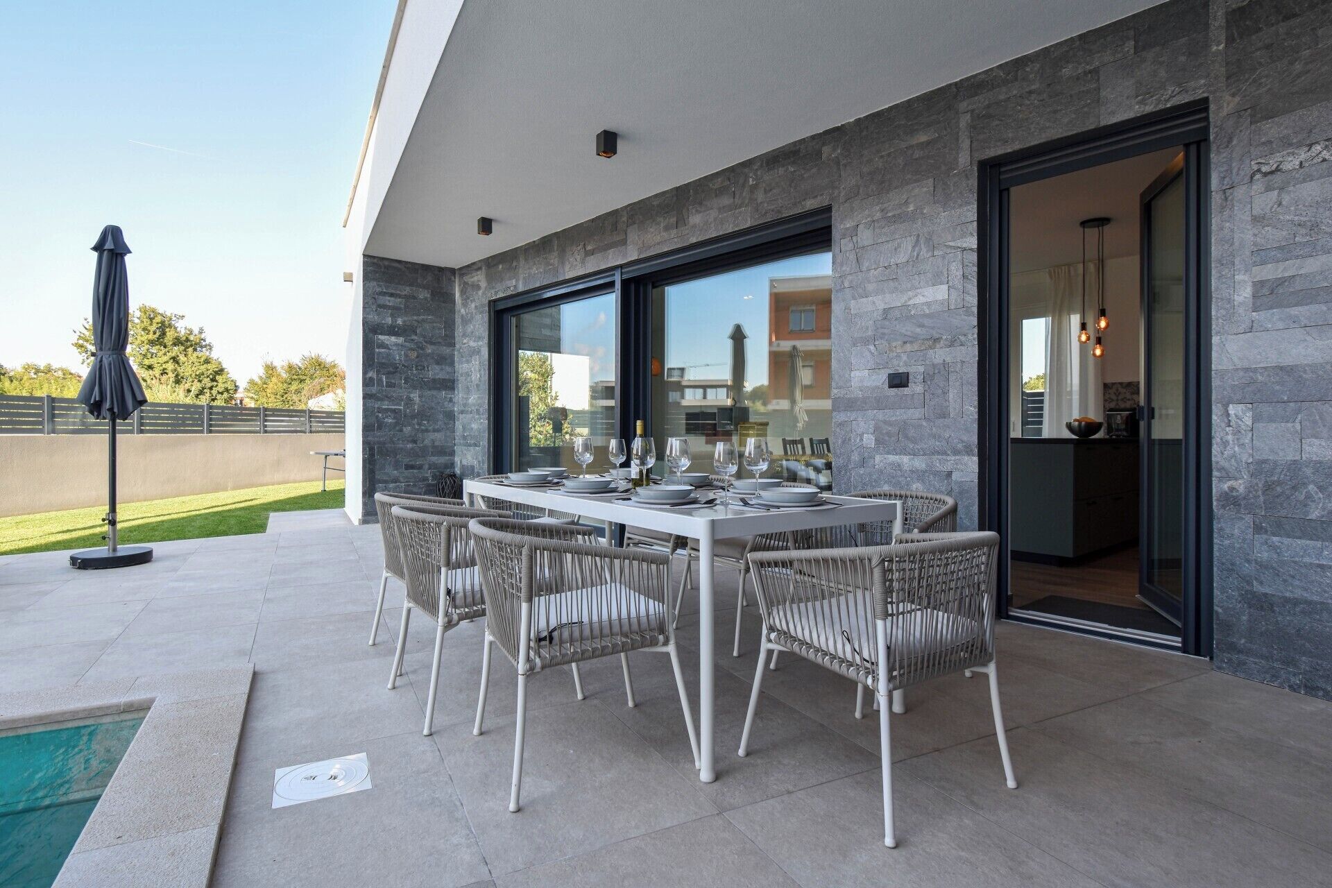 An outdoor dining area with a table set for six, surrounded by wicker chairs, against a modern building with stone walls and large windows.